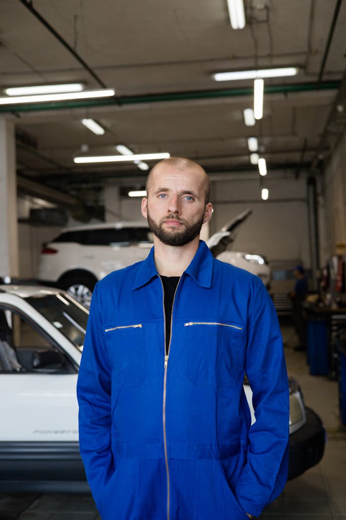 Caucasian mechanic in blue coverall standing in an auto repair shop with cars in the background.