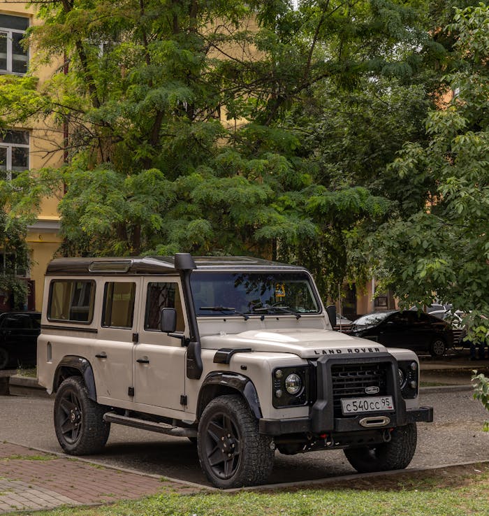 Vintage Land Rover Defender parked under green trees on a sunny day in the city.