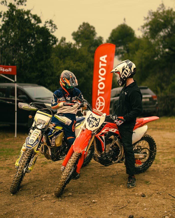 Two motocross riders with helmets prepare for a race in an outdoor rally scene.